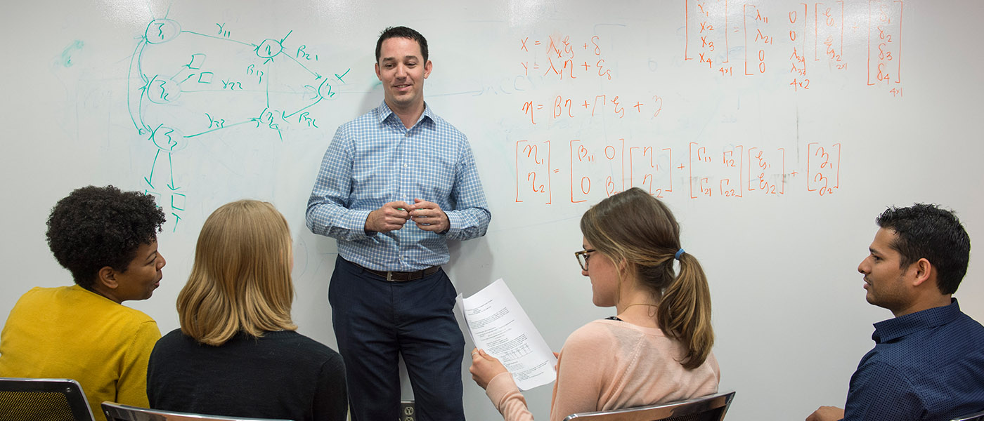 A professor in a plaid shirt stands in front of a whiteboard with green and red diagrams
